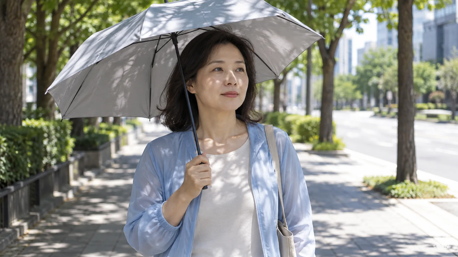 맑은 날 가로수 그늘이 진 도심 인도에서 하늘색 긴소매 겉옷을 입은 40대 여성이 무늬 없는 회색 양산을 쓰고 걷고 있다.